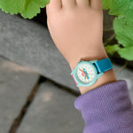 Ein Kind, das eine Cool Time Kids Armbanduhr - The Cool Rainbow Cloud von Cool Time und einen lila Ärmel trägt, steht neben grünen Blättern und grauen Steinen.