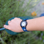 Eine Kinderhand mit einer Cool Time Kids Armbanduhr - The Cool New Soccer von Cool Time hält eine grüne Gießkanne im Freien.
