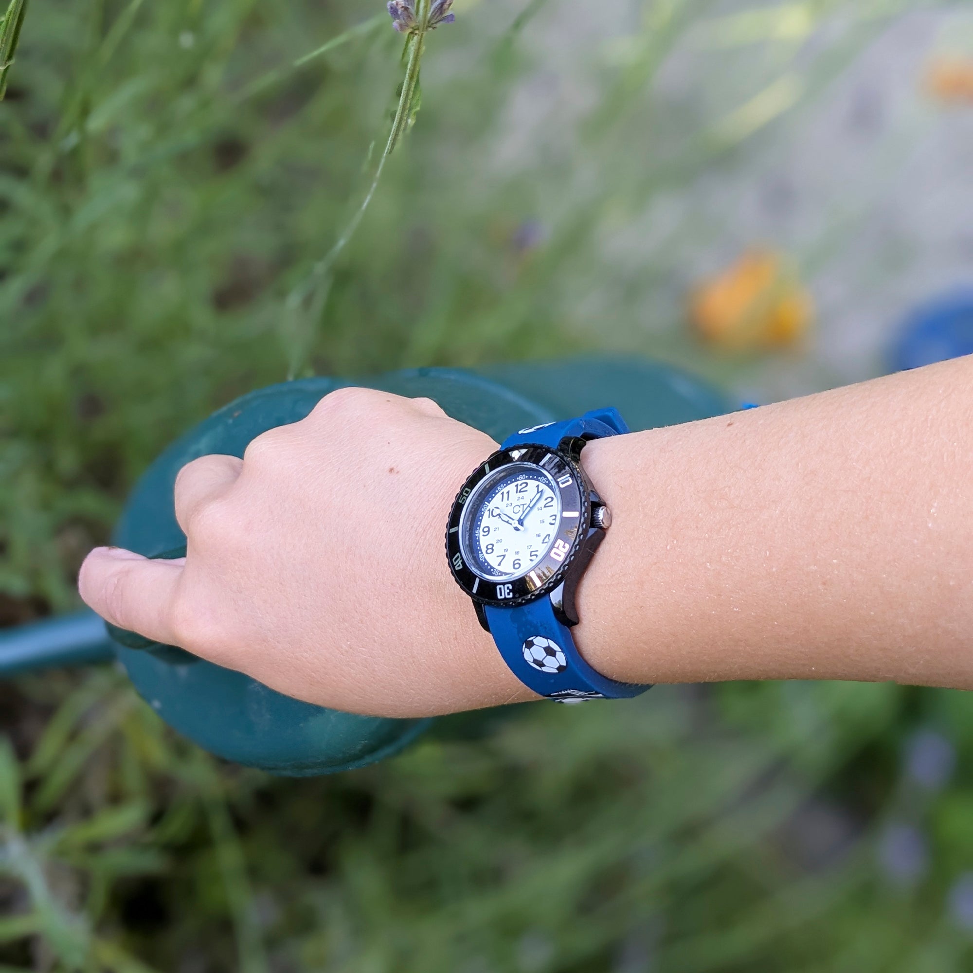 Eine Kinderhand mit einer Cool Time Kids Armbanduhr - The Cool New Soccer von Cool Time hält eine grüne Gießkanne im Freien.