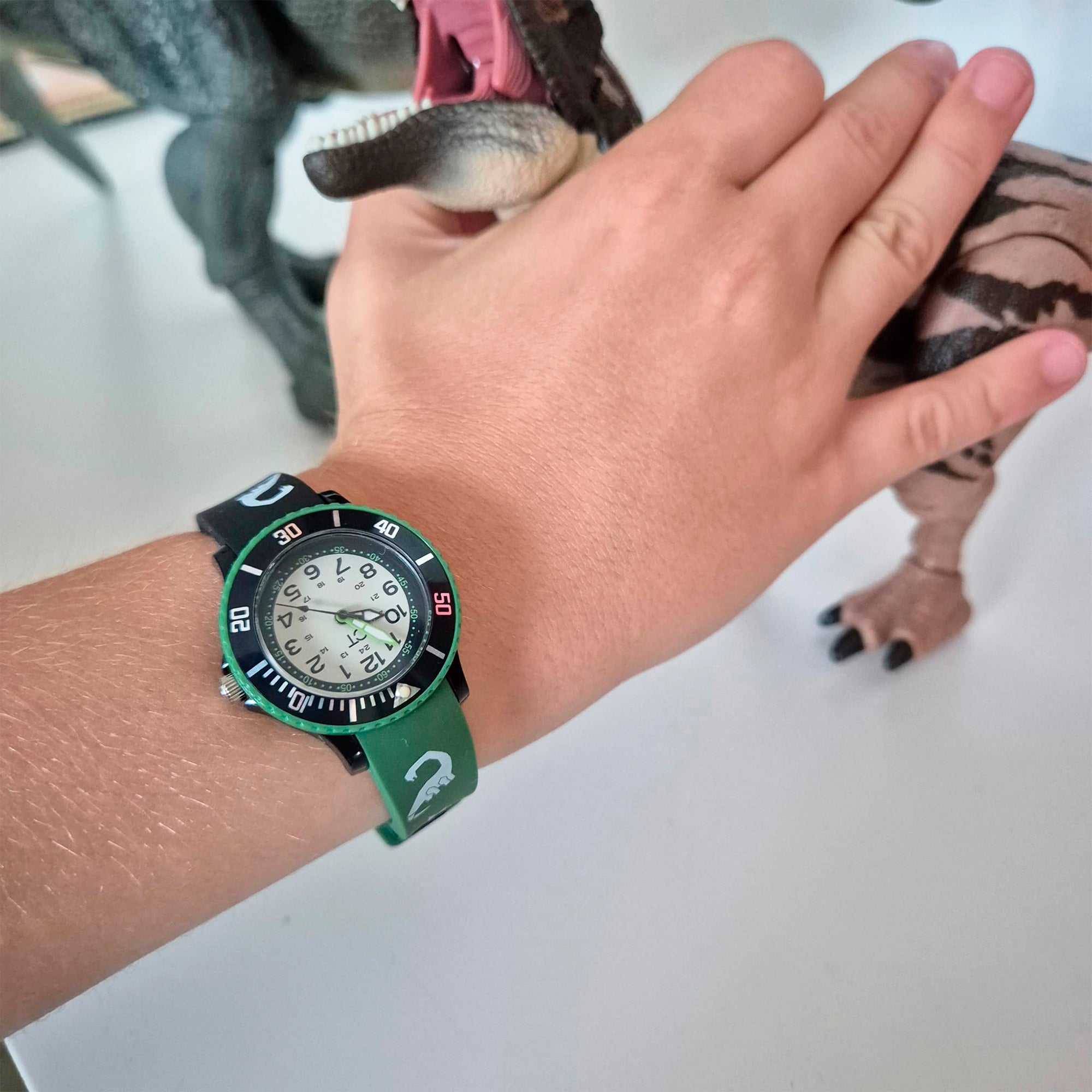 Eine Hand, die die Cool Time Kids Armbanduhr - The Cool New Dino von Cool Time trägt, hält ein Dinosaurier-Spielzeug mit offenem Mund.
