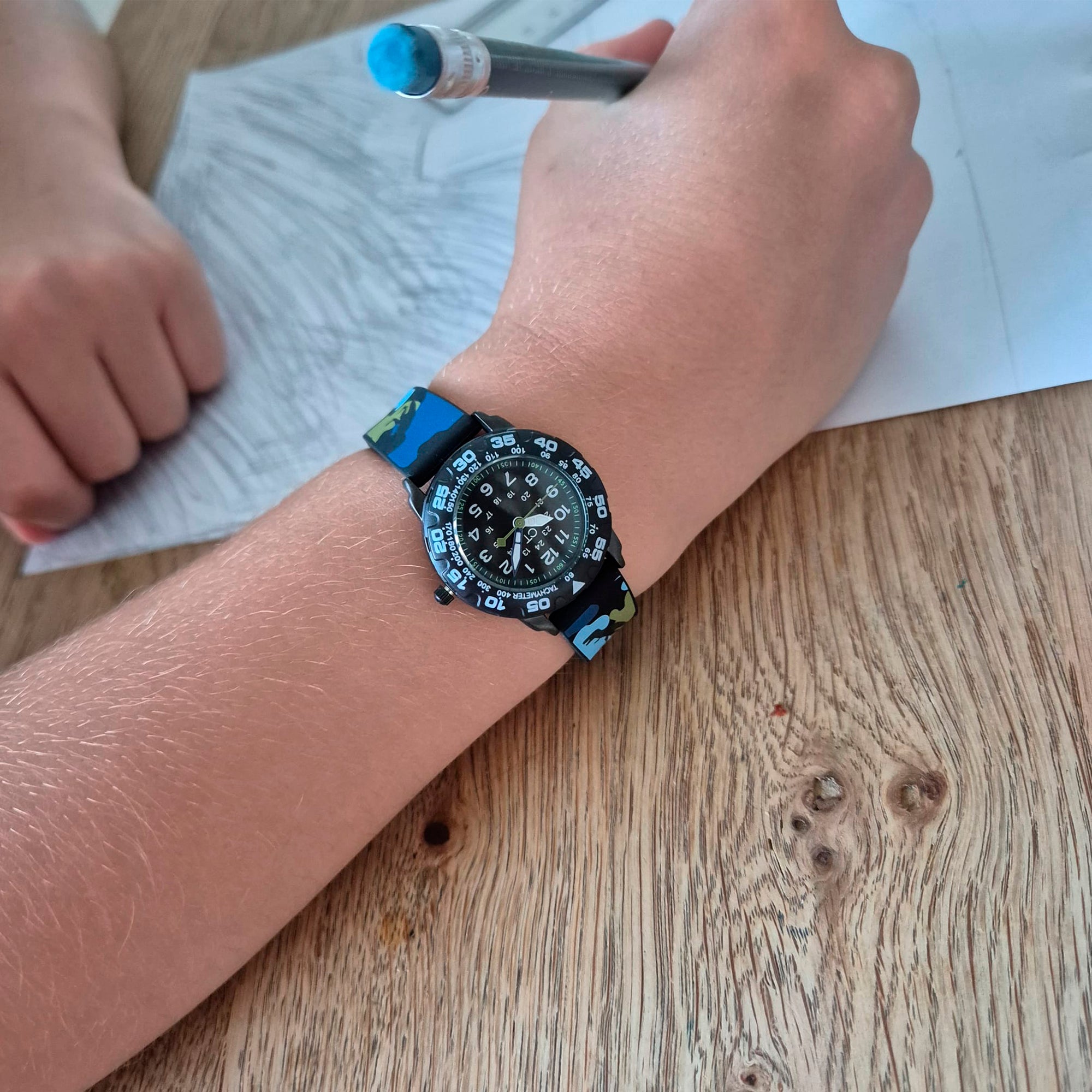 Eine Kinderhand, die die Cool Time Kids Armbanduhr - The Cool Dino Camouflage von Cool Time trägt, zeichnet mit einem Bleistift auf weißem Papier an einem Holztisch.