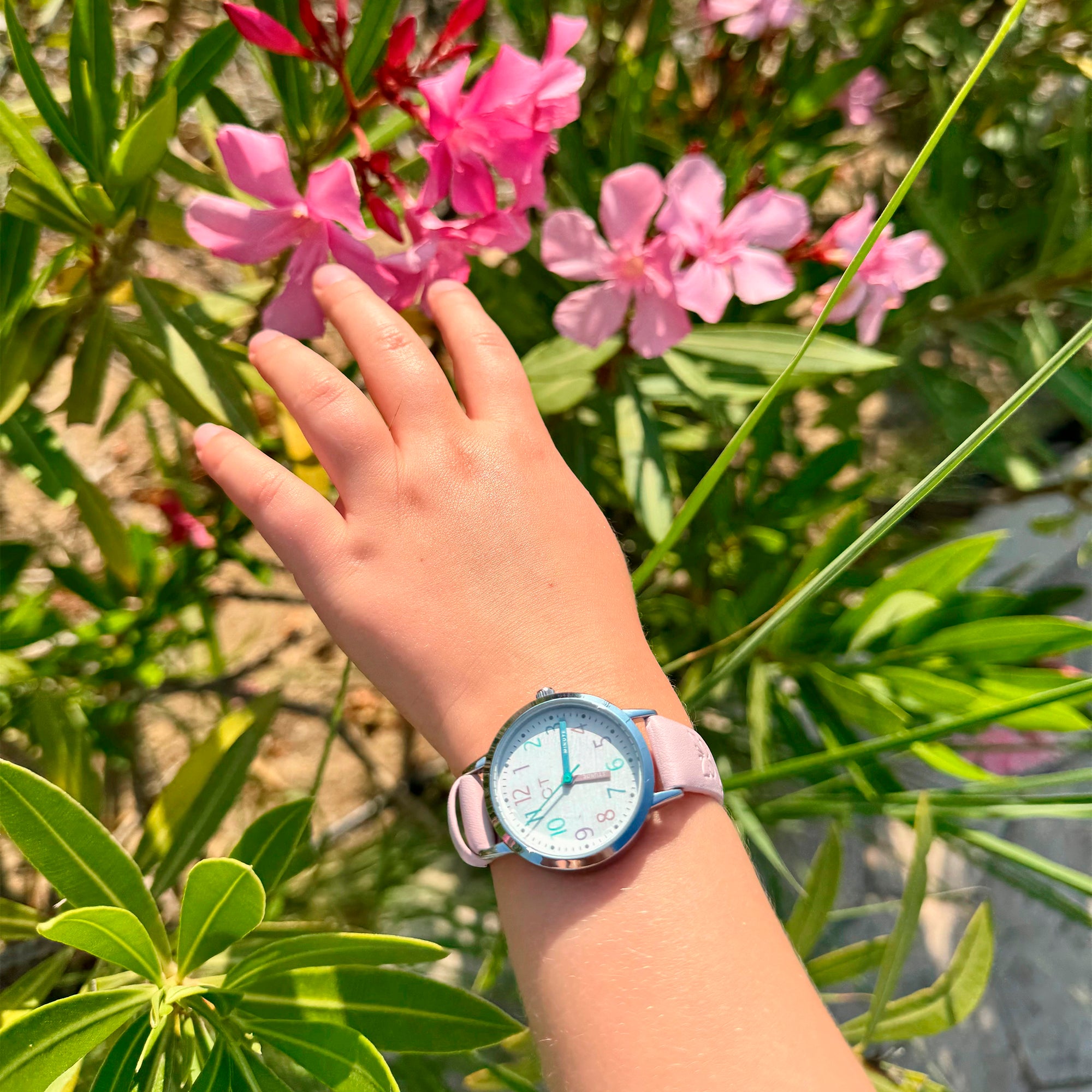 Eine Kinderhand mit einer Cool Time Kids Armbanduhr - The Cool Pink Pastel Butterfly streckt sich nach rosa Blumen im hellen Sonnenlicht.