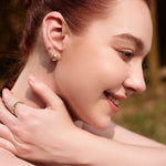 Eine Frau lächelt im Freien, sie trägt Ania Haie Ohrringe - Gold Kiss Hoops von Ania Haie und einen passenden Ring, ihre Hand berührt sanft ihren Hals.