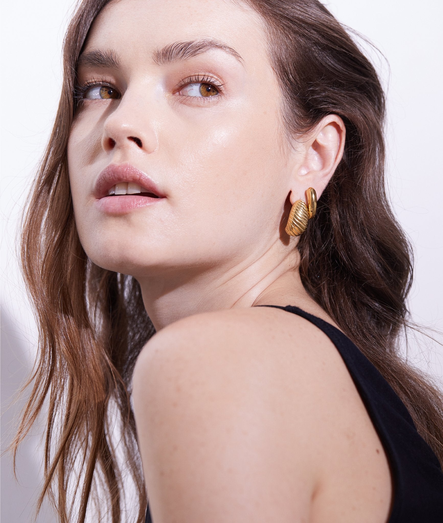 Model goldene Ohrringe der Marke „Echtzeit“. Sie blickt über die Schulter nach hinten, während weiches Licht ihr Gesicht und die Ohrringe betont.