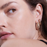 Nahaufnahme eines Frauengesichts mit braunen Augen, das den goldenen, muschelförmigen Creolen-Ohrring "The Organic Drop" von Echtzeit und ein schwarzes Trägertop trägt - ein raffiniertes Stück von Damen Schmuck.