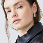 Eine Frau in schwarzer Lederjacke trägt Echtzeit Creole - The Organic Drop Ohrringe von Echtzeit und blickt selbstbewusst in die Kamera vor einem hellen Hintergrund.