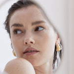 Frau mit nassem Haar und goldenen Echtzeit Creole - Die Organic Drop Ohrringe von Echtzeit schauen zur Seite, reflektiert in einem abstrakten Spiegelrahmen - ein schickes Damen Accessoire.