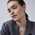 Eine Frau in einem grauen Blazer trägt die Echtzeit Creole - Like Diamonds Silberohrringe und eine mehrlagige Halskette von Echtzeit und blickt selbstbewusst in die Kamera.