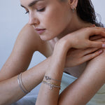 Eine Frau mit nassem Haar trägt silberne Ohrringe und legt das Echtzeit Armband - The Sleek One von Echtzeit übereinander, wobei sie ihr Kinn für den perfekten Lagenlook auf ihre Hände stützt.