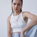 Eine Frau mit nassen Haaren, bekleidet mit einem weißen Tanktop und Jeans, trägt die Echtzeit Halskette - The Sleek One von Echtzeit und setzt den Lagenlook vor einem schlichten Hintergrund um.