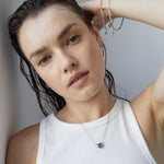 Eine Frau mit nassen Haaren in einem weißen ärmellosen Oberteil präsentiert eine Herz-Halskette und das Echtzeit Armband - The Sleek One von Echtzeit und kreiert einen schicken Lagen-Look, während sie in einem Haus posiert.