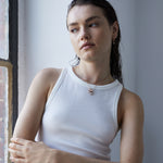 Frau mit nassen Haaren in einem weißen Tank-Top steht an einem Fenster und schaut nachdenklich zur Seite. Die Echtzeit Halskette - Like Diamonds von Echtzeit verleiht ihrem Look ein zeitloses, elegantes Accessoire.