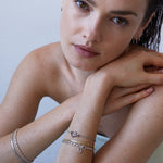 Eine Frau mit braunen Haaren stützt ihren Kopf auf ihre Arme und präsentiert das Echtzeit Armband - Like Diamonds von Echtzeit; gepaart mit Ohrringen wird dieser elegante Damen Schmuck Look durch silberne Details und ein Unendlichkeitszeichen-Motiv akzentuiert.