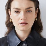 Eine Frau mit braunen Haaren trägt Echtzeit Creole - Modern Lines goldene Creolen von Echtzeit und eine schwarze Lederjacke und blickt in die Kamera - sie verkörpert zeitlose Eleganz und modernen Stil.