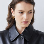 Frau mit gewelltem braunem Haar in schwarzer Lederjacke und Echtzeit Creole - Modern Lines von Echtzeit, die zeitlose Eleganz verkörpert. Sie blickt zur Seite, ihre modernen Ohrringe verleihen ihrem Stil einen Hauch von Raffinesse.