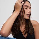 Frau mit nassem, gewelltem Haar in einem schwarzen Top berührt sanft ihren Kopf, sie trägt das Echtzeit Armband - Love Letters von Echtzeit mit lila Band und Buchstabenanhängern.