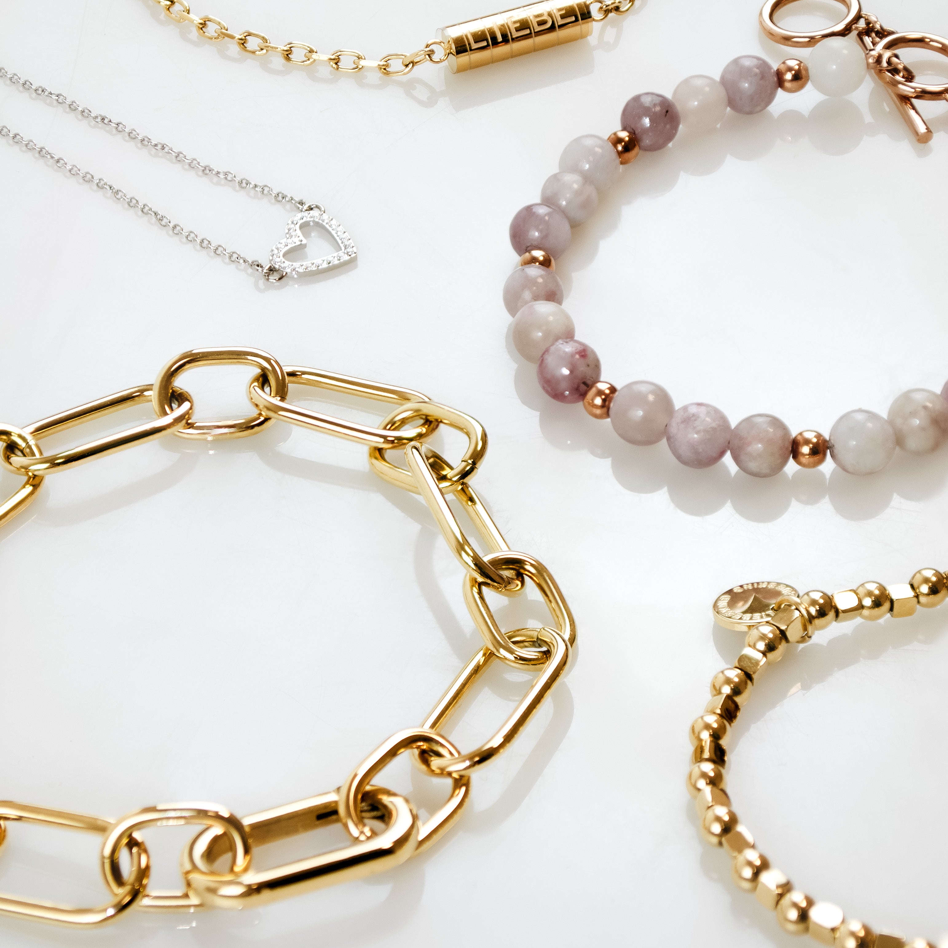 Flatlay mit verschiedenen Damenarmbändern und einer filigranen Herzkette in Gold- und Silber-Optik. Zu sehen sind ein grobgliedriges Kettenarmband, ein Perlenarmband mit Naturstein-Optik, ein Kugelarmband sowie ein feines Armband – arrangiert auf hellem Hintergrund.