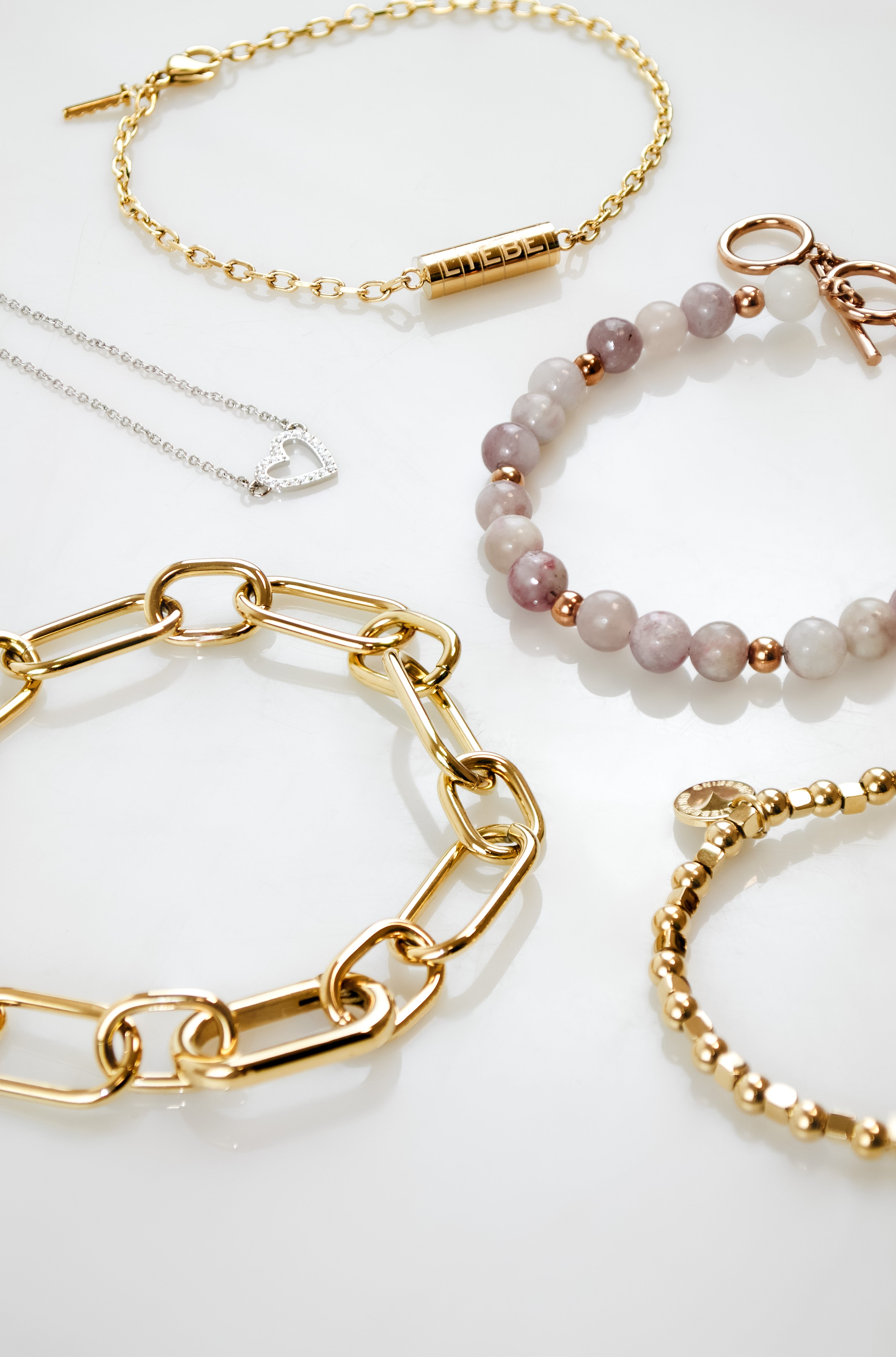 Flatlay mit verschiedenen Damenarmbändern und einer filigranen Herzkette in Gold- und Silber-Optik. Zu sehen sind ein grobgliedriges Kettenarmband, ein Perlenarmband mit Naturstein-Optik, ein Kugelarmband sowie ein feines Armband – arrangiert auf hellem Hintergrund.