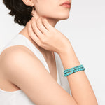 Eine Frau in einem ärmellosen weißen Top berührt ihren Hals und trägt das Liebeskind Berlin Beads - The SmallTurquoise Armband.
