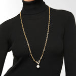 Eine Person trägt die Liebeskind Berlin Halskette - The Long Pearl, eine lange Goldkette mit einem einzelnen Perlenanhänger, über einem schwarzen Rollkragenpullover.