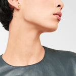 Frau trägt einen kleinen goldenen LIEBESKIND BERLIN Ohrstecker - The Geometric von Liebeskind Berlin, gepaart mit einem dunklen Ledertop und vom Hals aufwärts gezeigt.