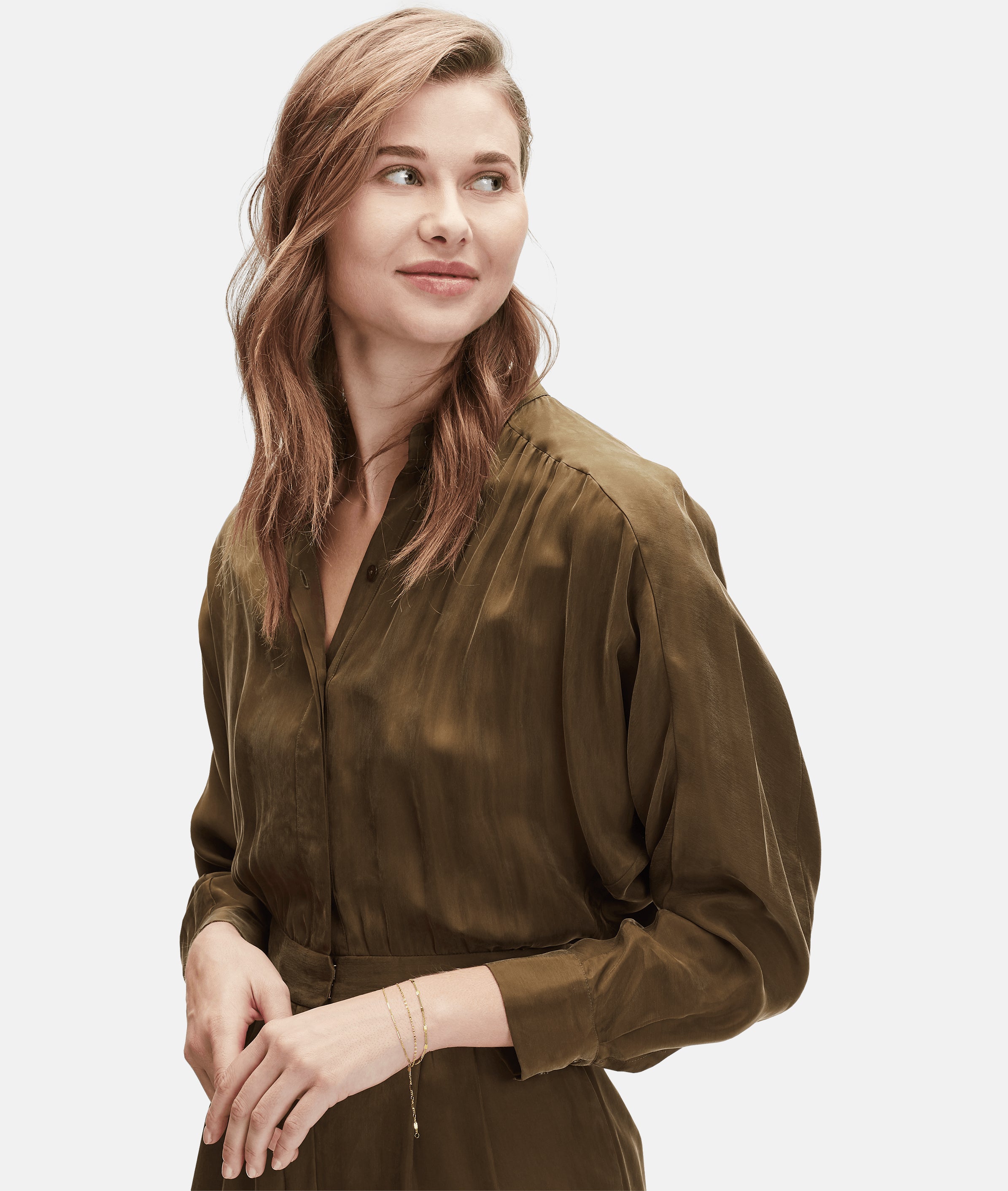 Frau mit gewelltem braunem Haar und olivgrüner Bluse, zur Seite lächelnd, mit einer goldenen Uhr und dem Liebeskind Berlin Gold Everyday Layering Set ausgestattet.