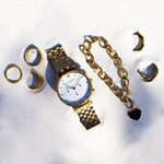 Die LIEBESKIND BERLIN Armbanduhr - Modern Chronograph von Liebeskind Berlin sowie goldene Ringe, ein Armband und Ohrringe liegen auf weißem Sand im strahlenden Sonnenlicht.