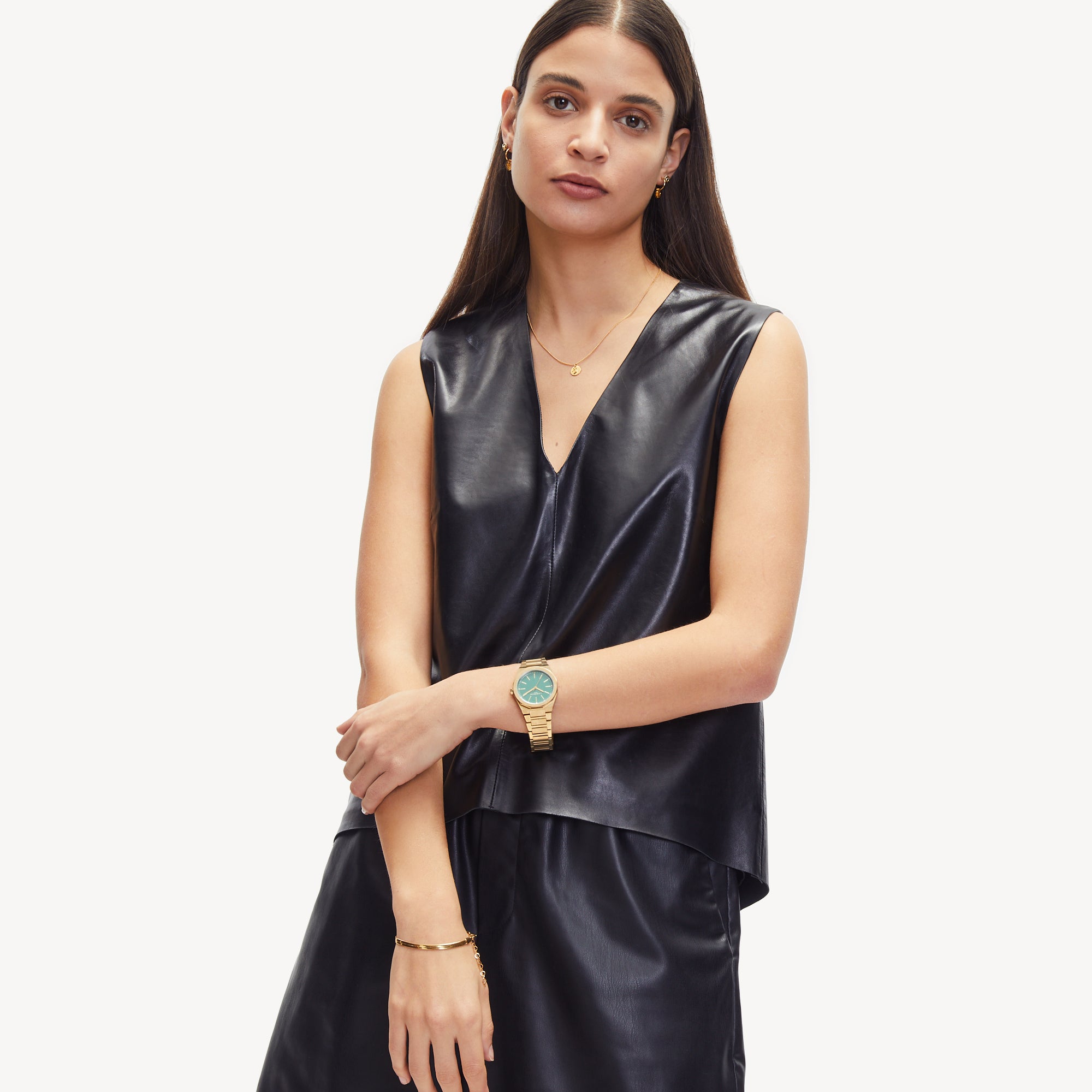 Eine Frau in einem ärmellosen schwarzen Ledertop und Rock, ausgestattet mit Goldschmuck und dem Liebeskind Berlin Set - Golden Basic, steht vor einem weißen Hintergrund.