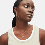 Frau mit geflochtenem Haar trägt ein beigefarbenes Tanktop, geometrische Ohrringe und die Liebeskind Berlin LIEBESKIND BERLIN Halskette - Swinging Icon mit passendem Anhänger.