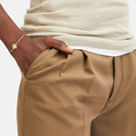 Eine Person, die eine hellbraune Hose und ein cremefarbenes, geripptes Oberteil trägt, mit einer Hand in der Tasche, ausgestattet mit dem goldenen LIEBESKIND BERLIN Armband - Swinging Icon von Liebeskind Berlin.