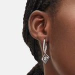 Nahaufnahme eines Liebeskind Berlin Swinging Icon Ohrhänger aus Silber mit einem quadratischen Logomania-Anhänger, getragen von einer Person mit geflochtenem Haar.