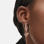 Nahaufnahme des Ohrs einer Frau, die den goldenen Ohrhänger - Swinging Icon von Liebeskind Berlin mit einem quadratischen Anhänger vor einem schlichten Hintergrund trägt.