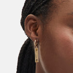 Nahaufnahme einer Person mit geflochtenem Haar, die einen goldenen rechteckigen LIEBESKIND BERLIN Ohrhänger - Secure Tag Icon Designer-Ohrring von Liebeskind Berlin trägt.