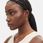 Frau mit geflochtenem Haar in einem niedrigen Pferdeschwanz, trägt goldenen Schmuck, darunter die Liebeskind Berlin Halskette - The Love Lock und ein ärmelloses beigefarbenes Oberteil, Blick nach links.