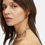 Frau mit braunen Haaren, die die LIEBESKIND BERLIN Halskette - The Secure Tag von Liebeskind Berlin und Ohrringe trägt und über ihre Schulter schaut.