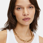 Frau in einem weißen Tank-Top trägt die LIEBESKIND BERLIN Halskette - The Secure Tag von Liebeskind Berlin, eine klobige Goldkette, und schaut in die Kamera.