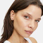 Frau mit braunen Haaren trägt die LIEBESKIND BERLIN Creole - The Secure Tag Ohrringe von Liebeskind Berlin und ein weißes Tanktop und schaut in die Kamera.