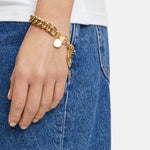 Eine Hand, die das Liebeskind Berlin Armband - The Organic, ein klobiges Edelstahlarmband mit einem Herzanhänger, trägt, ruht auf einer blauen Jeans und einem weißen Hemd.