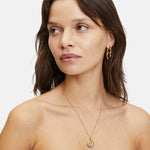 Die braunhaarige Frau trägt goldene Ohrringe, eine Liebeskind Berlin "The Organic Pendant" Halskette und ein schwarzes trägerloses Top.