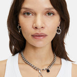 Frau mit schulterlangen braunen Haaren trägt ein weißes Tanktop und LIEBESKIND BERLIN The Organic Creole aus Edelstahl mit abnehmbarem Anhänger, dazu Reifohrringe von Liebeskind Berlin.