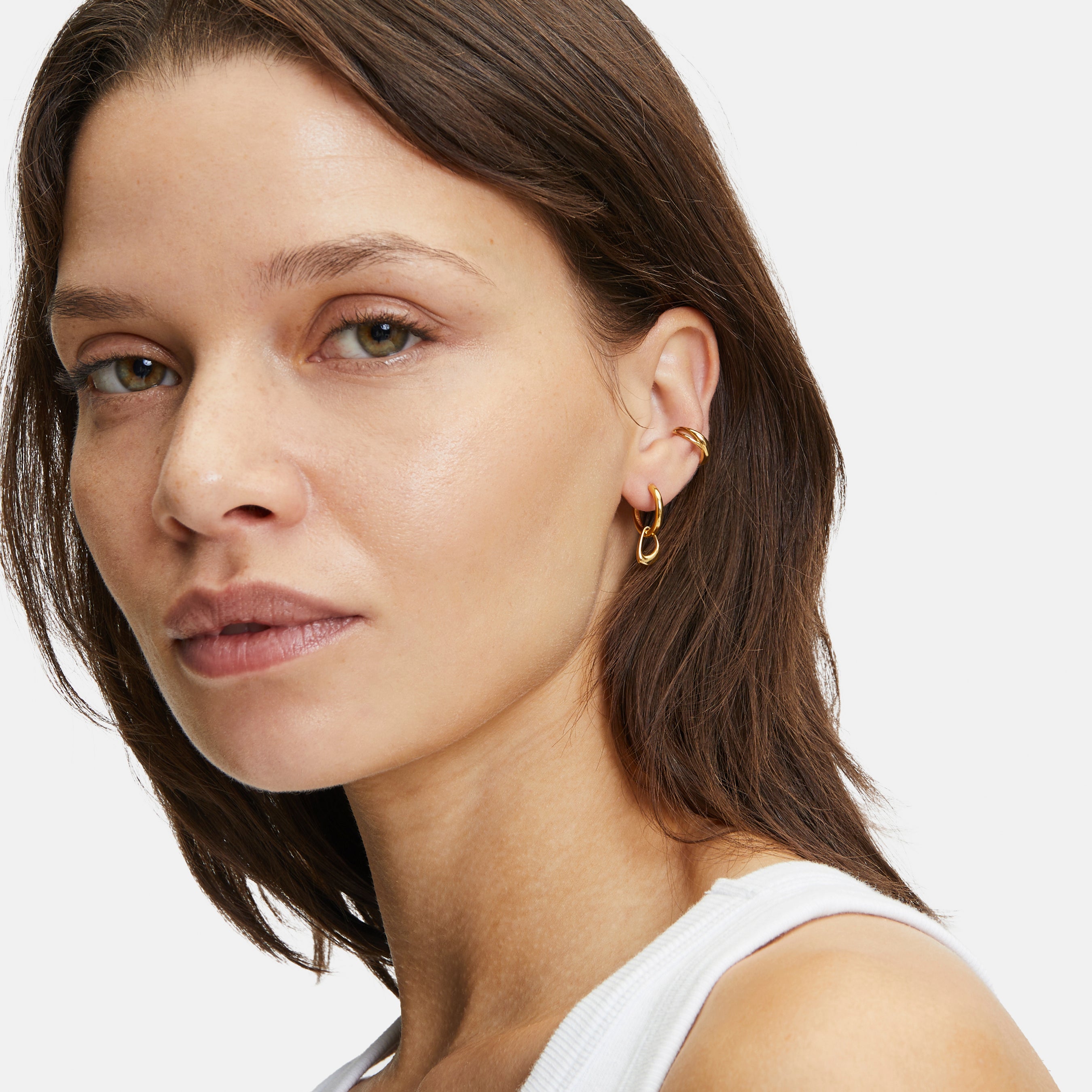 Frau mit glattem braunem Haar trägt Creole - The Organic Creole Creolen von Liebeskind Berlin und ein weißes Tanktop und schaut in die Kamera.