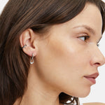 Eine Frau mit braunem Haar, im Profil, trägt drei silberne Ohrringe, darunter den LIEBESKIND BERLIN Ear Cuff - The Twisted von Liebeskind Berlin, zu einem weißen Oberteil.