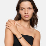 Frau mit gewellten braunen Haaren in einem trägerlosen schwarzen Oberteil posiert mit der Hand auf der Schulter und präsentiert mühelos den LIEBESKIND BERLIN Armband - Karabiner von Liebeskind Berlin, das Must-have Trend Accessoire Schmuck dieser Saison.