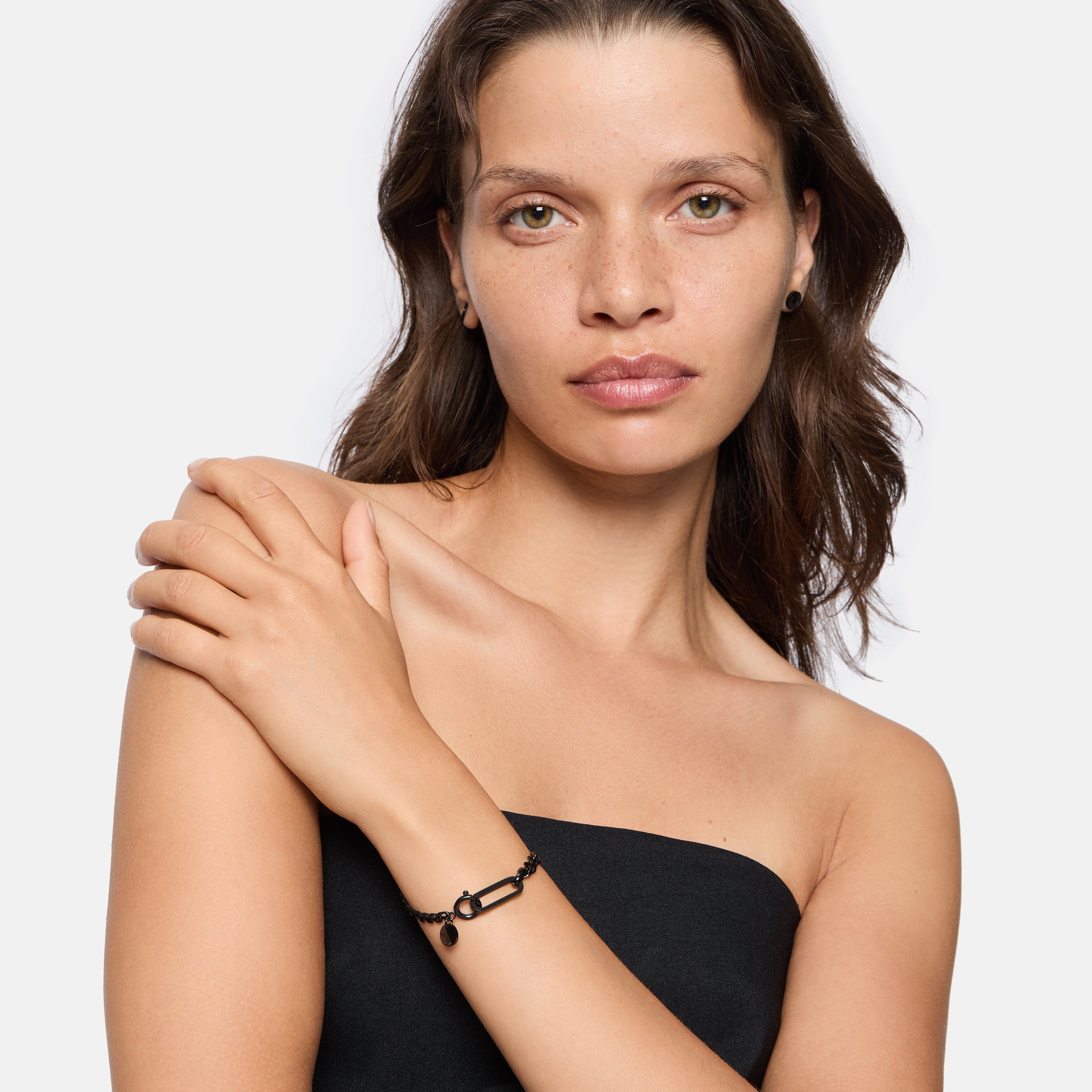 Frau mit gewellten braunen Haaren in einem trägerlosen schwarzen Oberteil posiert mit der Hand auf der Schulter und präsentiert mühelos den LIEBESKIND BERLIN Armband - Karabiner von Liebeskind Berlin, das Must-have Trend Accessoire Schmuck dieser Saison.