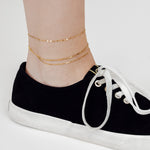 Eine Person trägt schwarze Sneakers mit weißen Schnürsenkeln und eine LIEBESKIND BERLIN Fußkette - Sparkling With Layers von Liebeskind Berlin als stylisches Accessoire am Knöchel.