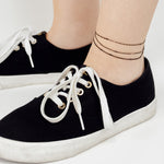 Nahaufnahme von Füßen in schwarzen Turnschuhen und der LIEBESKIND BERLIN Fußkette - Sparkling With Layers von Liebeskind Berlin, einem eleganten, mehrschichtigen Fußkettchen, vor hellem Hintergrund.