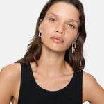 Frau mit gewellten braunen Haaren, die ein schwarzes Tanktop und LIEBESKIND BERLIN Creole - Sparkling Link Ohrringe von Liebeskind Berlin trägt, blickt in die Kamera.