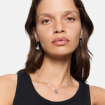 Frau mit braunen Haaren in einem schwarzen Tank-Top, trägt die LIEBESKIND BERLIN Creole - Urban Heart Ohrringe von Liebeskind Berlin und eine passende silberne Herz-Anhänger-Halskette.