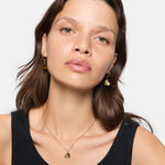 Eine Frau mit gewelltem braunem Haar, goldenen Ohrringen in Herzform und der LIEBESKIND BERLIN Halskette - Urban Heart von Liebeskind Berlin in einem schwarzen Tank-Top - eine Verkörperung von zeitloser Eleganz.