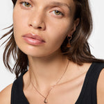 Frau mit schwarzem Oberteil, die Liebeskind Berlin Schmuck - eine goldene Halskette und LIEBESKIND BERLIN Creole - Urban Heart Ohrringe - trägt, blickt in die Kamera.